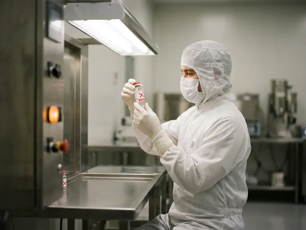 Pharmaceutical cleanroom technician in white gown and mask inspecting a rejected vial under fluorescent light with amber warning glow.