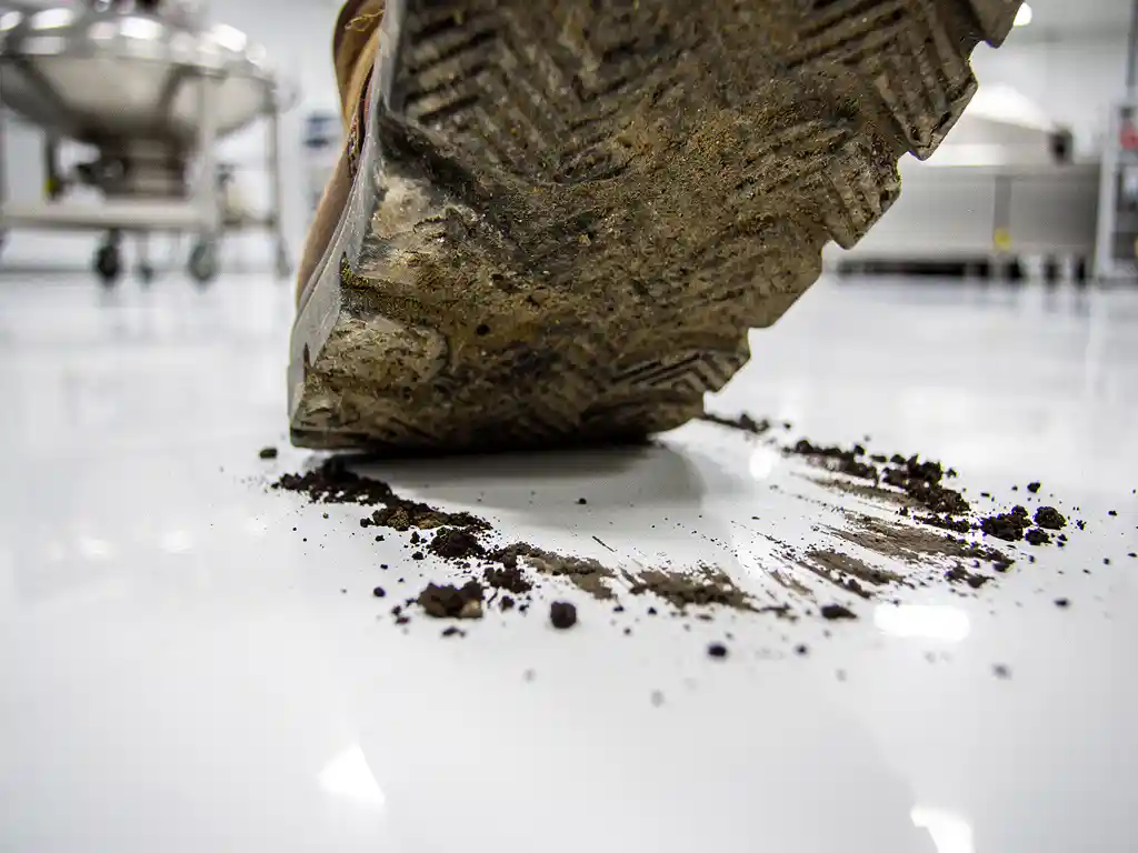 Schmutzige Arbeitsstiefel-Sohle hinterlässt Erdpartikel auf weißem Lebensmittelproduktionsboden, Edelstahlausrüstung im Hintergrund.